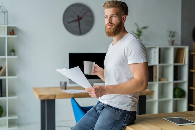 Barbu jeune homme d'affaires travaillant au bureau moderne. Homme portant un t-shirt blanc et prenant des notes sur les documents.