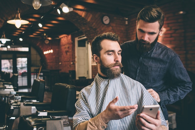 Un barbu montre au coiffeur une photo au téléphone. Le coiffeur regarde le téléphone avec une vue sérieuse.