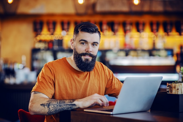 barbu tatoué jeune hipster caucasien assis dans un café et travaillant sur un projet indépendant.