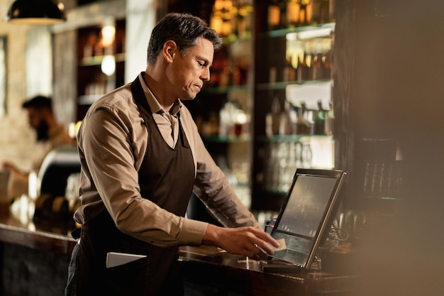 Barista adulte moyen vérifiant les commandes à la caisse enregistreuse tout en travaillant dans un pub.