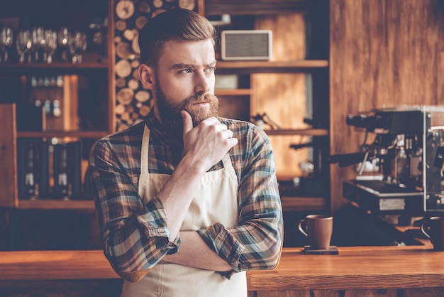 Photo barista attentionné. jeune homme barbu en tablier regardant loin et gardant la main sur le menton tout en se tenant au comptoir du bar