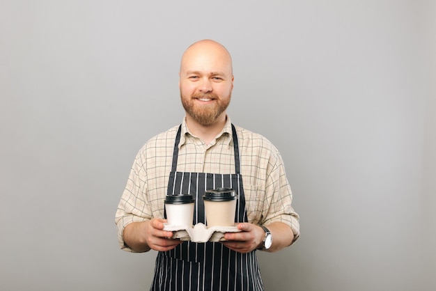 Barista souriant à la caméra alors qu'il tient deux gobelets en papier avec une boisson chaude dedans sur un mur gris