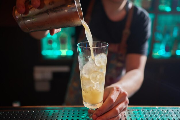 Barman en chemise et tablier faisant une boisson alcoolisée avec de la glace dans un verre à cocktail