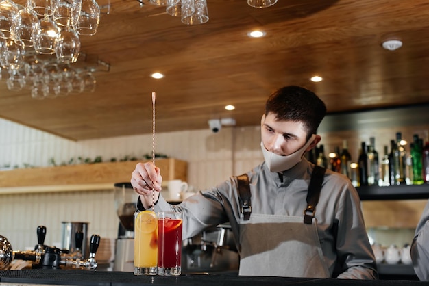 Photo un barman élégant portant un masque et un uniforme pendant une pandémie prépare des cocktails lors d'une fête. le travail des restaurants et des cafés pendant la pandémie.