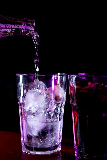 Le barman en gros plan verse de l'eau gazeuse dans un verre avec de la glace Mélanger un cocktail d'alcool dans un restaurant de pub de discothèque Éclairage rose atmosphérique sur fond noir