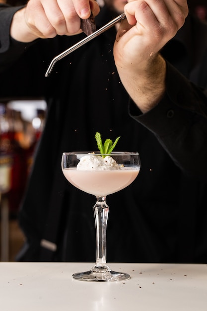 Le barman masculin frotte le chocolat avec une râpe sur un cocktail avec de la crème glacée et des Baileys dans un verre