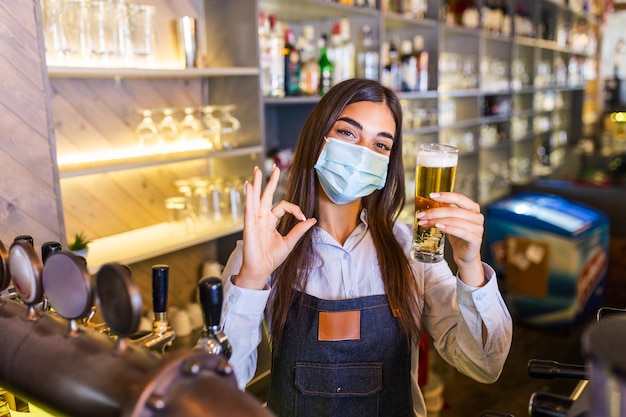 Barman avec un masque de protection du visage covid19 servant une bière au comptoir du bar pendant la pandémie de coronavirus montrant des étagères OK pleines de bouteilles avec de l'alcool en arrière-plan