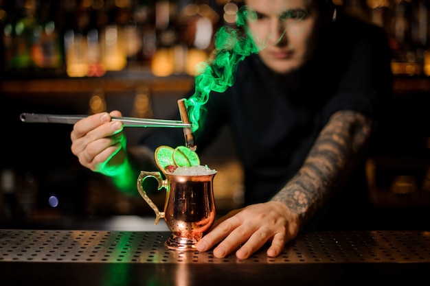 Barman professionnel s'ajoutant à un cocktail dans le verre de tonnelier avec une cannelle fumée aromatique orange séchée avec des pincettes sur le comptoir du bar