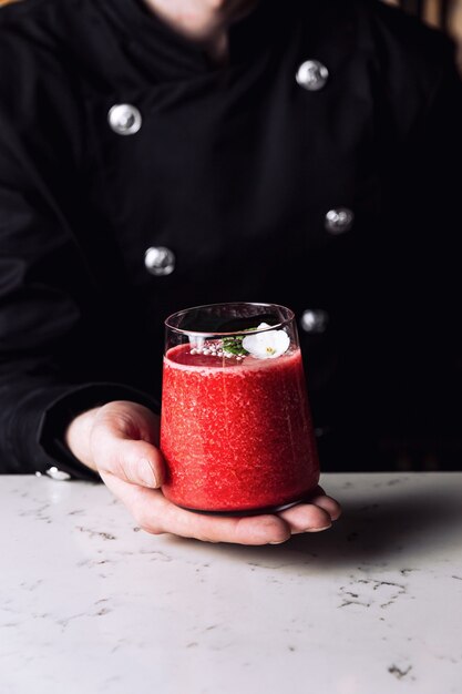 Barman tenant un verre de cocktail rafraîchissant à la fraise et à la banane garni d'une fleur comestible, table en marbre.