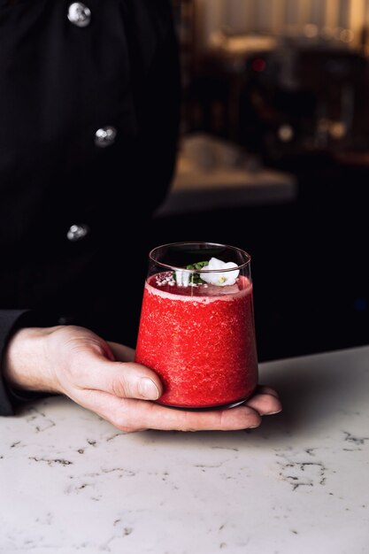 Barman tenant un verre de cocktail rafraîchissant à la fraise et à la banane garni d'une fleur comestible, table en marbre.