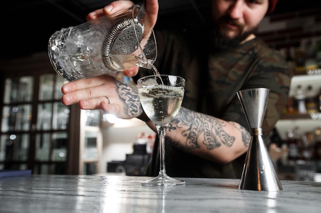 Barman versant un alcool de la tasse de mesure en verre à travers la passoire au cocktail sur le comptoir du bar à l'arrière-plan flou