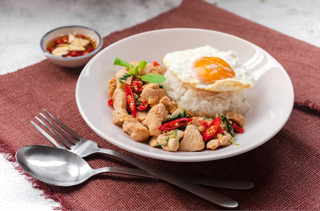 Basilic sauté garni de riz avec du poulet et un œuf au plat sur une assiette blanche servant sur une nappe brune avec sauce de poisson Thai street food