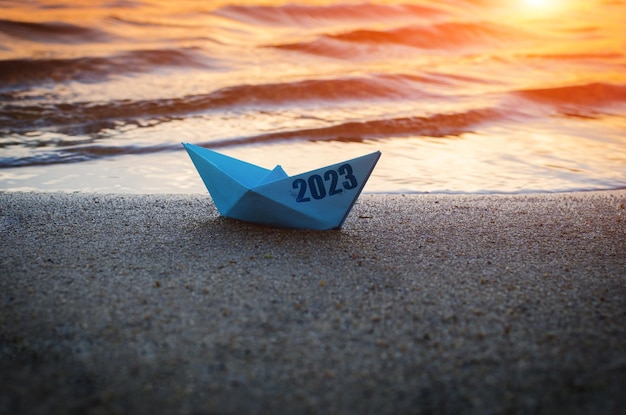 Bateau en papier bleu avec inscription 2023 sur la plage de sable contre le coucher du soleil