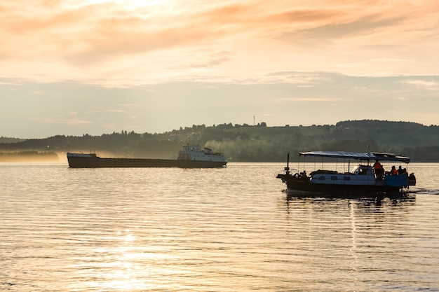 Un bateau à passagers et une barge cargo descendent une large rivière dans la lumière du soir