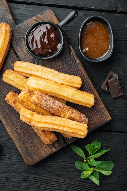 Bâtonnets de churros traditionnels avec ensemble de poudre de cannelle et de sucre, sur fond de table en bois noir, vue de dessus à plat