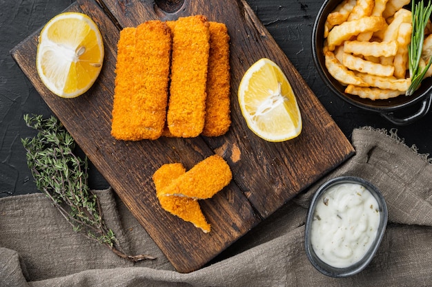 Bâtonnets de poisson avec pommes de terre frites sur planche à découper en bois sur fond noir vue de dessus mise à plat