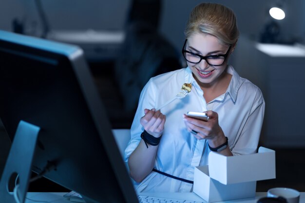 Bavarder avec des amis . Bonne secrétaire agréable et agréable assis dans le bureau et manger de la restauration rapide tout en exprimant la positivité et en tenant le téléphone