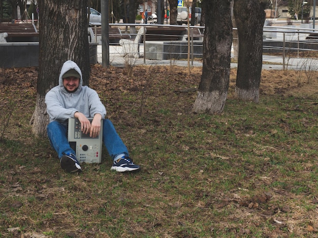 Un beatmaker hiphop est assis sur l'herbe dans un parc sous un arbre tenant une boîte à rythmes à l'ancienne des années 90 Instrument de musique numérique rétro pour producteurs et beatmakers de hip hop