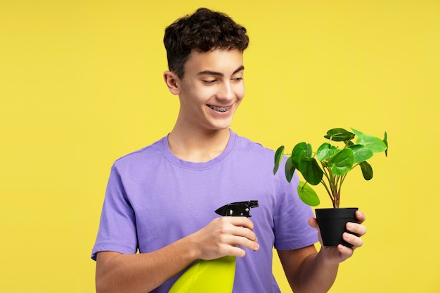Un beau adolescent souriant avec des appareils orthopédiques tenant un pot de fleurs le pulvérisant isolé sur un fond jaune