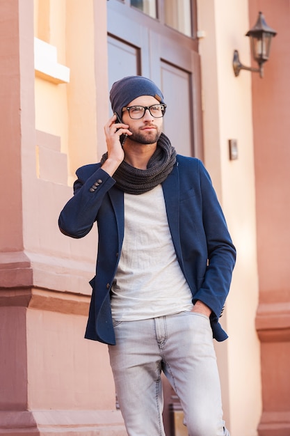 Beau au téléphone. Beau jeune homme en vêtements décontractés intelligents parlant au téléphone portable tout en se tenant devant l'entrée de la maison et en tenant une tasse de café