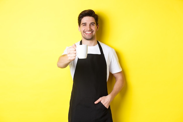 Beau barista en tablier noir vous donnant une tasse de café et souriant, debout sur fond jaune.