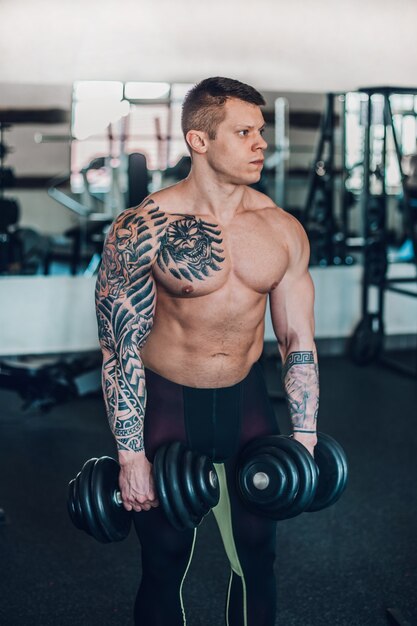 Beau bodybuilder masculin avec des haltères debout dans la salle de fitness