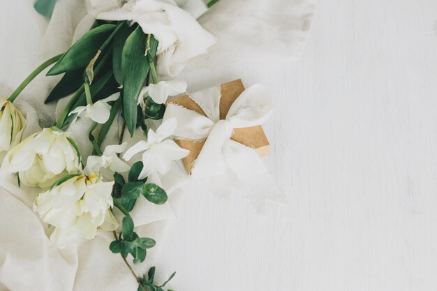 Beau bouquet blanc avec boîte-cadeau sur une table en bois rustique à plat Bonne fête des mères Tulipes printanières élégantes et jonquilles sur tissu doux et présent Journée de la femme Espace pour le texte