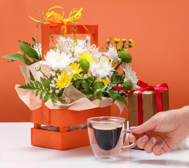 Beau bouquet de fleurs jaunes et blanches dans le panier en bois, un coffret cadeau et une main de femme avec une tasse de café.