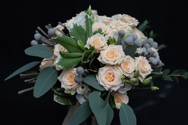 Beau bouquet de mariage composé de rose crème touffue, d’eucalyptus, de Brunei, de Pittosporum et de Lisianthus sur un mur noir.