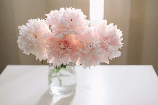 Beau bouquet rose clair fraîchement coupé de pivoines roses dans un vase en verre sur une table blanche près d'une fenêtre
