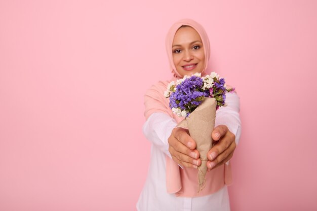 Un beau bouquet simple de fleurs sauvages de prairie dans des tons violets, enveloppé dans du papier kraft brun dans les mains d'une jolie femme musulmane souriante portant un hijab. Espace de copie de fond rose isolé