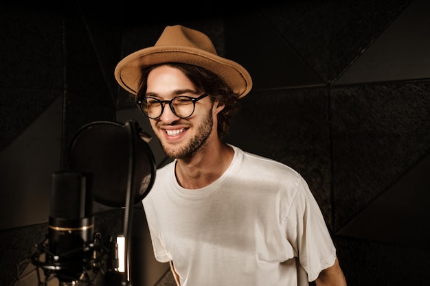 Beau chanteur masculin souriant chantant joyeusement en enregistrant une nouvelle chanson pour un album de musique dans un studio moderne. Musicien attrayant travaillant dans un studio d'enregistrement