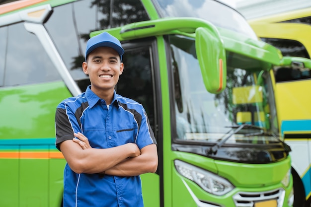 Un beau chauffeur de bus en uniforme et chapeau sourit mains croisées avec l'arrière-plan du bus