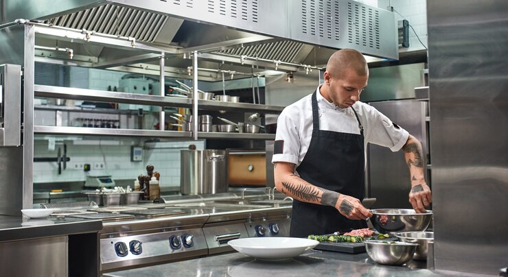 Photo beau chef professionnel avec des tatouages mélangeant une salade dans un bol en métal en se tenant debout dans une cuisine
