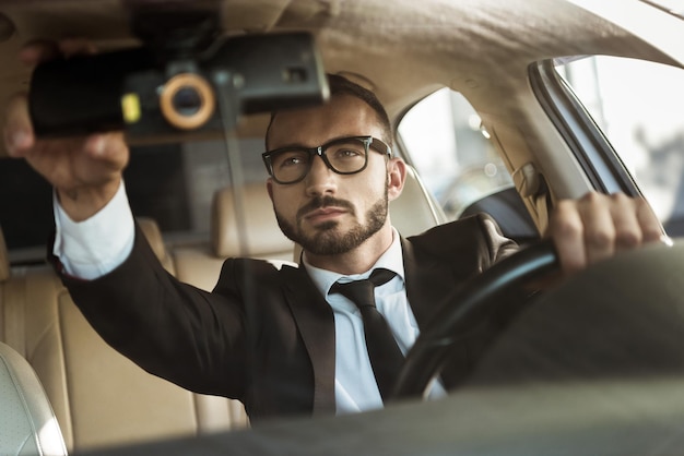 Beau conducteur en costume conduisant une voiture et fixant un miroir