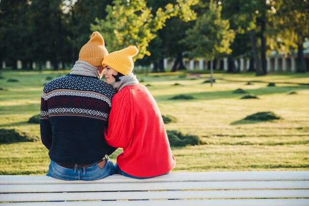 Beau couple assis sur un banc, porte des vêtements chauds et des chapeaux tricotés, s'embrassent