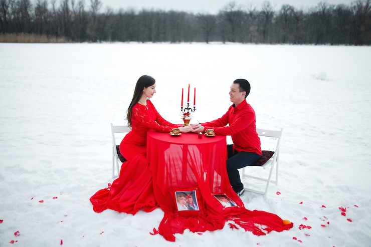 Photo beau couple assis à une table avec un chandelier sur fond de neige en vêtements rouges