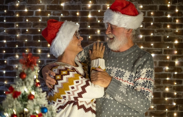 Beau couple caucasien senior fête Noël à la maison embrassé un couple âgé en pull et chapeaux de père noël se regardant dans les yeux