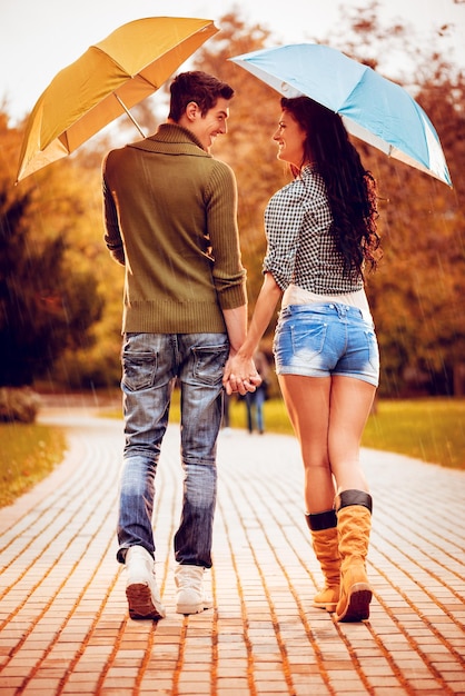 Beau couple charmant avec des parapluies profitant de la marche sous la pluie à travers le parc aux couleurs de l'automne. Vue arrière.