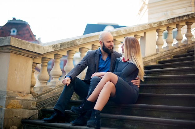 Un beau couple élégant de jeune femme et homme senior avec longue barbe noire assis embrassant près les uns des autres en plein air dans la rue d'automne dans les escaliers journée ensoleillée, photo horizontale