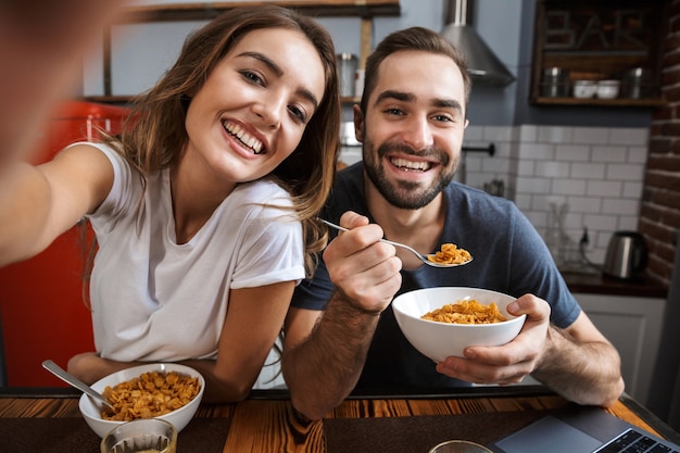 Beau couple gai prenant son petit déjeuner à la cuisine, prenant un selfie