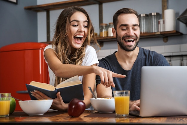 Beau couple gai prenant son petit déjeuner dans la cuisine, à l'aide d'un ordinateur portable, en lisant un livre