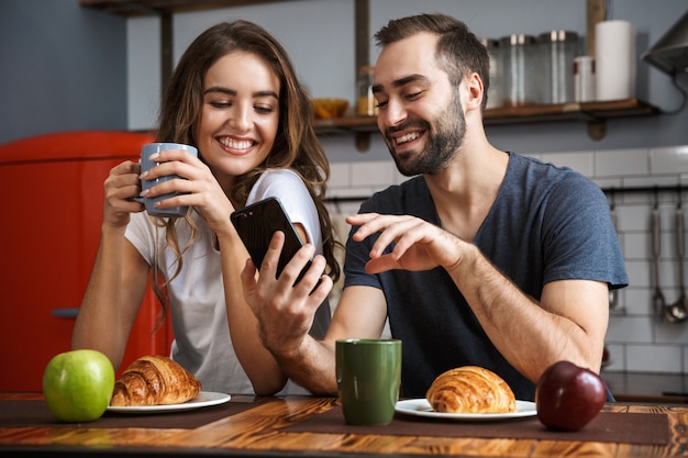 Beau couple gai prenant son petit déjeuner dans la cuisine, à l'aide de téléphone mobile