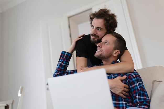 Beau couple gay riant ensemble assis dans leur salon à la maison Deux jeunes amants romantiques s'amusant à surfer sur Internet à l'intérieur Jeune couple gay vivant ensemble