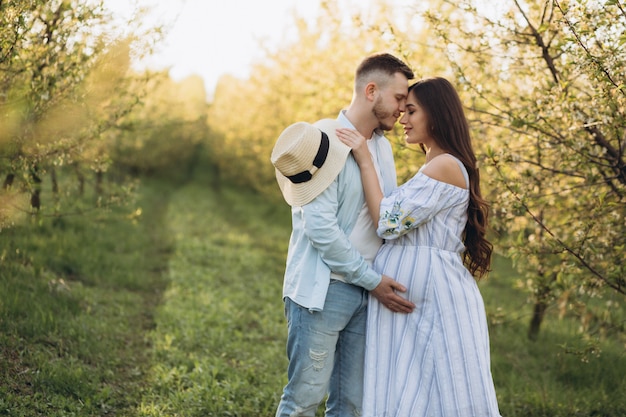 Beau couple, homme et femme enceinte en fleurs de couronne sur la tête, étreignant dans le jardin d'arbre de printemps. Maman heureuse attend bébé!