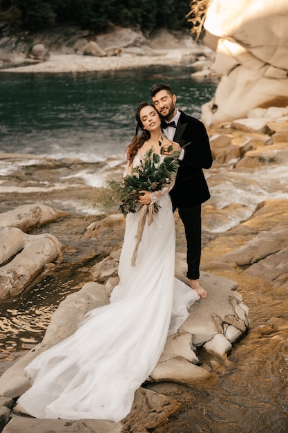 Beau couple de mariage étreint tendrement sur le fond d'une rivière de montagne, longue robe blanche de la mariée.