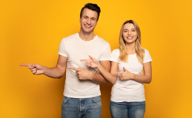 Un beau couple pose sur un fond jaune, portant des t-shirts blancs, pointant vers la gauche avec leurs mains et souriant à la caméra.