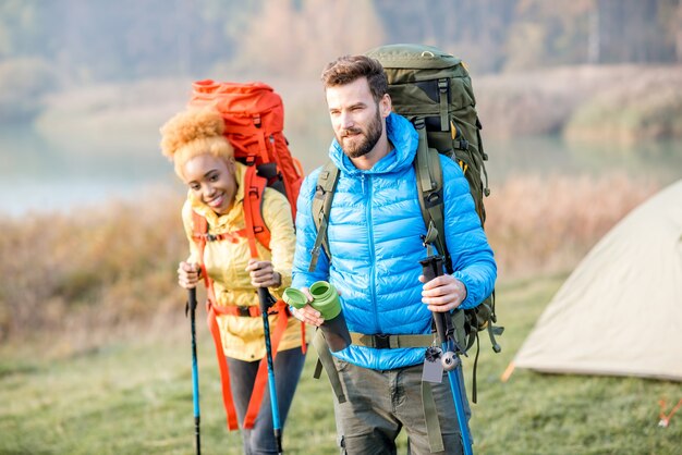 Beau couple en randonnée avec des sacs à dos colorés sur la pelouse verte avec tente en arrière-plan