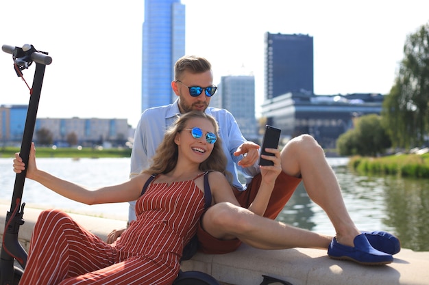 Beau couple s'amusant à conduire un scooter électrique, faisant une pause dans la conduite, assis au bord de la rivière, prenant un selfie,