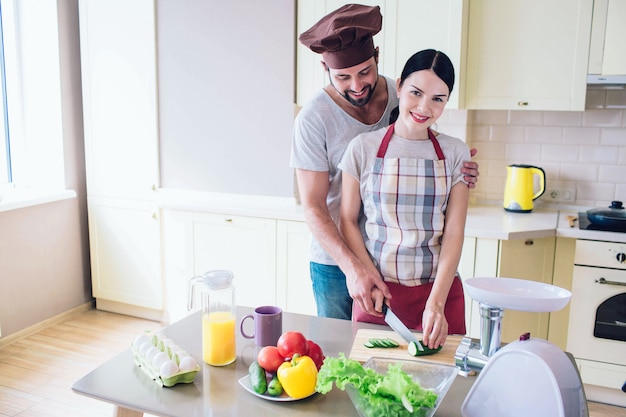 Beau couple se tient à la cuisine et wokr ensemble. La fille regarde et sourit. Elle tient un couteau dans une main et du concombre dans l'autre. Guy l'aide à couper des légumes.